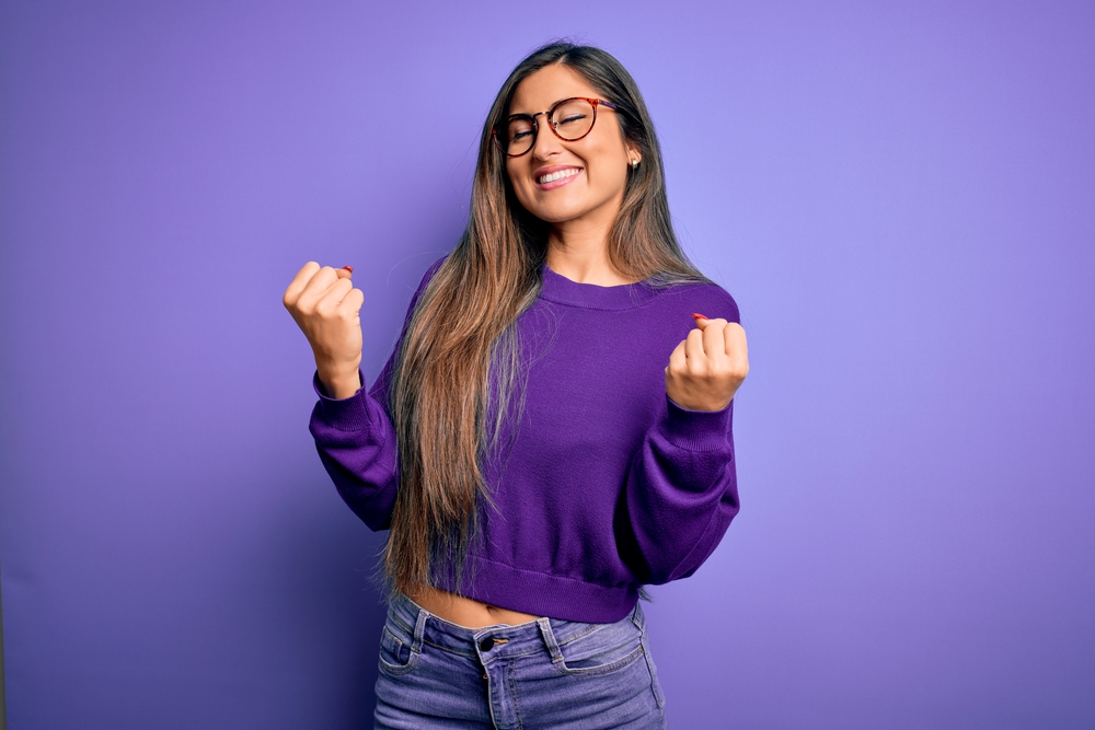 A joyful young woman in a purple sweater and glasses, clenching her fists in excitement over improved communication efficiency, standing against a purple background.