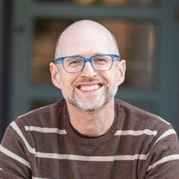 A smiling person with glasses and a beard wearing a brown striped sweater sits in front of a blurred background. A smiling person with glasses and a beard wearing a brown striped sweater sits in front of a blurred background.