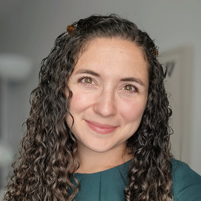 A woman with long curly brown hair and a green top smiles at the camera against a neutral background.