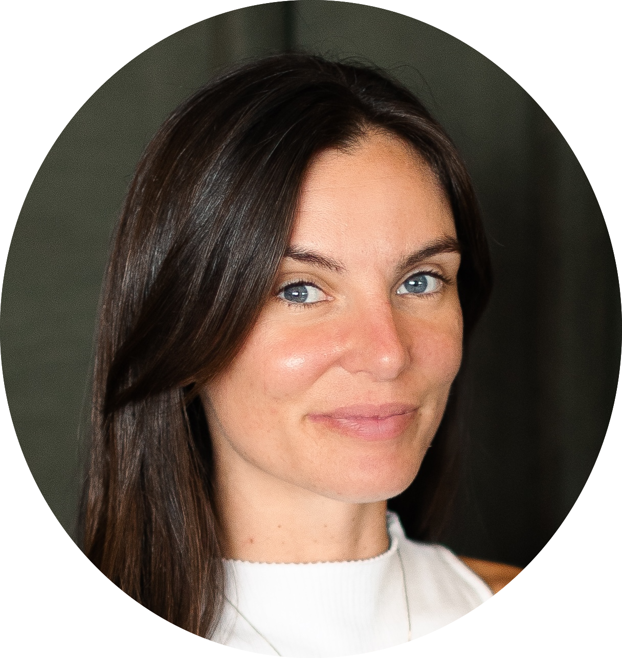 A woman with long brown hair and blue eyes, wearing a sleeveless white top, smiles slightly while looking at the camera.