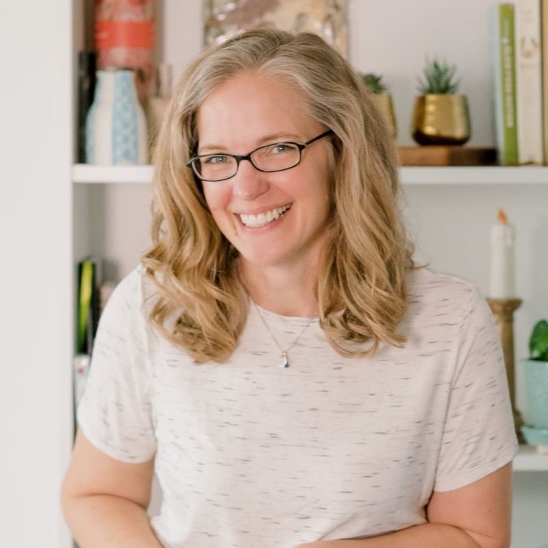 Woman with blonde hair and glasses smiles at the camera, wearing a light-colored shirt, standing in front of a bookshelf with various decor items.