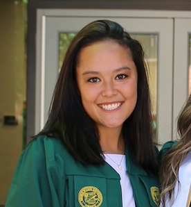 A woman wearing a green graduation gown and white shirt smiles at the camera, standing in front of glass doors.