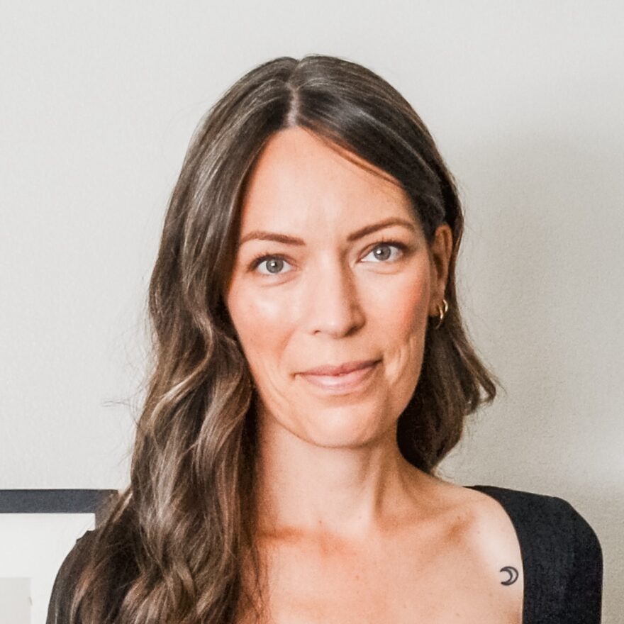 A woman with long wavy brown hair wearing a black top stands indoors next to a framed artwork and a light switch.