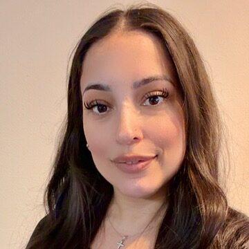 A woman with long, wavy dark hair wearing a black top and a necklace with a cross pendant, standing against a plain light-colored background.